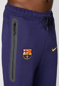 Marineblauwe joggingbroek met een zwart ritsvak, getextureerde grijze accenten en een geborduurd FC Barcelona-logo in geel en rood. Elastische taille met trekkoord.