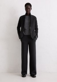 Black pinstriped blazer with a tailored fit, paired with a gray crew neck sweater and wide-leg pinstriped trousers. Flat black shoes.