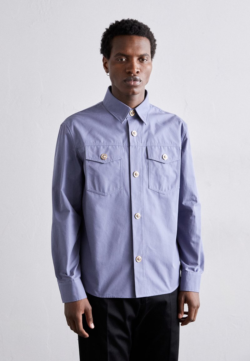Camisa de botones de color púrpura claro que presenta dos bolsillos frontales con detalles de botones dorados, mangas largas y un cuello estructurado.