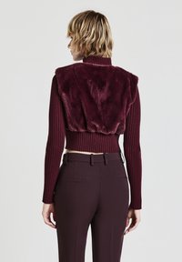 Bordeauxfarbener kurzer Pullover mit gerippten Ärmeln und Kunstpelzdetail auf dem Rücken. Kombiniert mit eng anliegenden dunklen Hosen, die eine schlanke Silhouette zeigen.