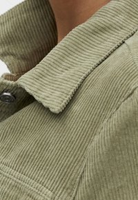 Veste en velours côtelé vert olive avec un col à revers et un détail à boutons. Le tissu présente une texture côtelée qui accentue son apparence tactile.