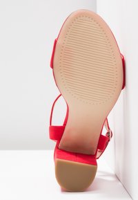 Red suede high-heeled sandal with a textured beige rubber sole, ankle strap, and buckle detail. Flat heel and sleek design.