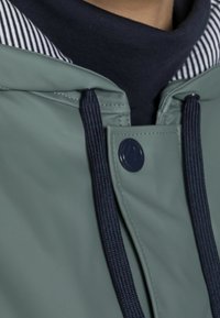 Vihreä sadeviitta, jossa on tummansiniset narut ja napinappi, päällä musta-valkoinen raidallinen paita ja tumman sininen poolopaita.