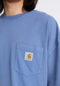 Lysblå kortærmet t-shirt lavet af bomuld, med en brystlomme, der har et gult og hvidt Carhartt logo. Blød tekstur, afslappet pasform.