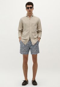 Chemise en lin beige à boutons associée à un short bleu à motifs. Le mannequin porte des lunettes de soleil noires et des mocassins marron, avec les mains dans les poches.