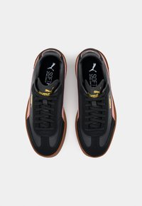 Baskets noires à surface texturée, accents roses, lacets plats, et semelle en caoutchouc marron. Présentent le logo Puma en or.