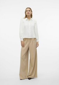 Blusa bianca a maniche lunghe con dettagli a ruffles sulle spalle, abbinata a pantaloni beige a vita alta e a gamba larga, caratterizzati da una texture liscia.