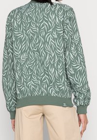 Zielona bluza z wzorem liści, ściągacze na rękawach i u dołu, wykonana z miękkiego materiału, z małą naszywką logo w dolnym lewym rogu.