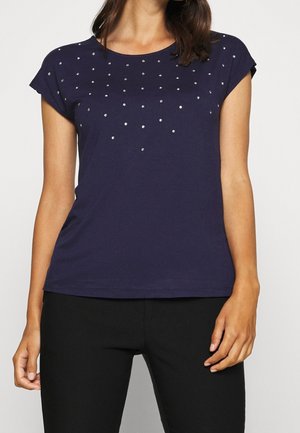 Vrouw die een marineblauw topje met korte mouwen draagt, met kleine zilveren studs verspreid over de borst, gecombineerd met een zwarte broek.