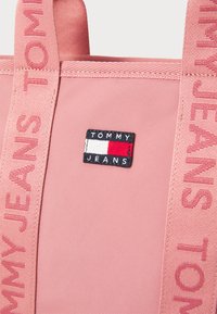 Roze stoffen tote bag met een gestructureerde afwerking, zijhandvatten en een patchlogo met rode, witte en blauwe elementen.