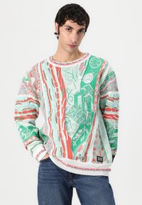 Un pull en tricot coloré présentant un mélange de vert, rouge et blanc avec des motifs abstraits. Encolure et poignets côtelés, avec des accents texturés.