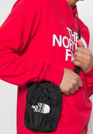 Sac pochon noir à cordon avec logo, en matériau léger, tenu dans une main, porté aux côtés d'un hoodie rouge avec un grand texte blanc.