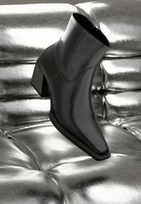 Bottine noire en cuir brillant, avec un bout pointu, un talon épais et un design sans couture, reposant sur une surface argentée réfléchissante.