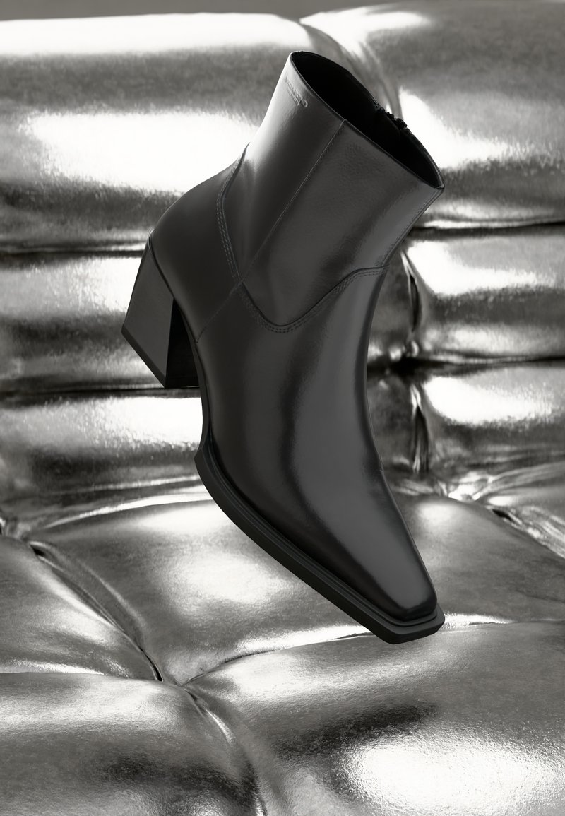 Bottine noire en cuir brillant, avec un bout pointu, un talon épais et un design sans couture, reposant sur une surface argentée réfléchissante.