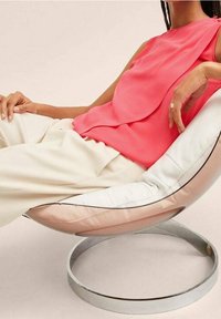 Korallenfarbenes ärmelloses Oberteil mit drapiertem Design, kombiniert mit hellbeigen Hosen. Weiche Texturen und ein runder Stuhl mit metallischem Gestell und Polsterung.