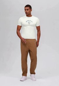 White short-sleeve t-shirt with "BROOKLYN NY" print, paired with tan trousers and white sneakers. The shirt has a round neckline.