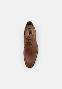 Zapato de vestir de cuero marrón con textura suave, punta afilada y cordones negros, con un cuello acolchado y el logotipo de la marca en el interior.