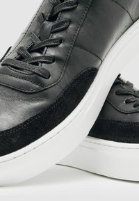 Schwarze Leder- und Wildledersneaker mit runder Zehe, flacher weißer Gummisohle und Schnürdesign. Sichtbare Steppnähte an den Seitenpaneelen.