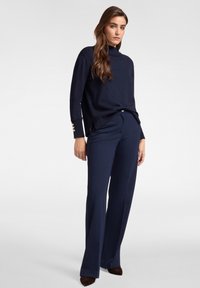Navyblauw gebreid sweater met hoge hals en zijsplitsingen, gecombineerd met brede navy broeken. De sweater heeft gouden knoopdetails op de mouwen.