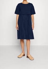 Marineblaues Baumwollkleid mit einem kurz geschnittenen Oberteil und lockerer Passform, kombiniert mit beigefarbenen, geflochtenen Clogs mit niedrigem Holzabsatz.