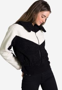 Chaqueta de cremallera en blanco y negro con un diseño en forma de chevrón, hecha de material suave, con cuello y cintura elástica, acompañada de pantalones negros.