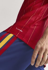 Homme portant un maillot de sport rouge avec des coutures jaunes marqué "Climacool" et un short bleu avec des rayures rouges et jaunes, main reposant le long du corps.