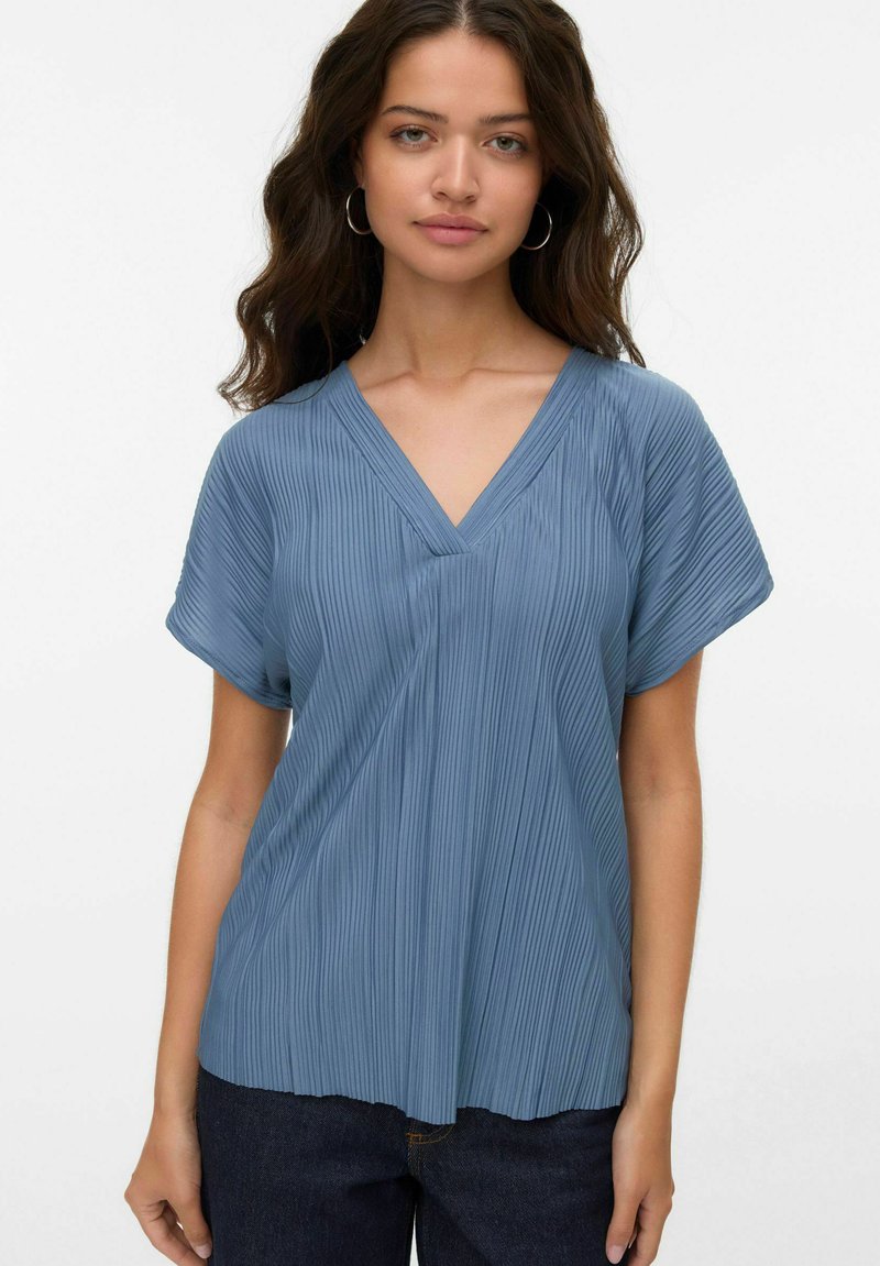 Giovane donna con lunghi capelli ondulati che indossa orecchini a cerchio in argento, una blusa blu a pieghe con scollatura a V e maniche corte, e jeans scuri, in piedi su uno sfondo bianco.