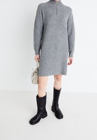 Personne portant une robe pull en tricot gris avec une courte fermeture éclair, des bottes noires jusqu'aux genoux et tenant un sac à main beige clair.