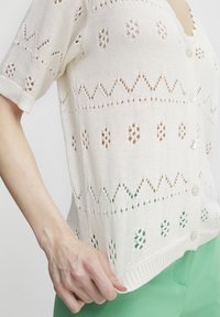 Knitted white cardigan with openwork patterns, short sleeves, and button closure. Features geometric designs along the body and hem.
