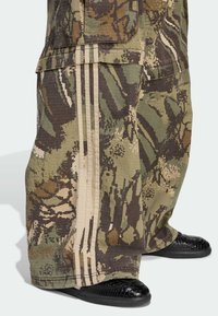 Pantalones cargo de camuflaje en verde, marrón y beige con rayas laterales prominentes y tejido texturizado. Combinados con calzado negro texturizado.