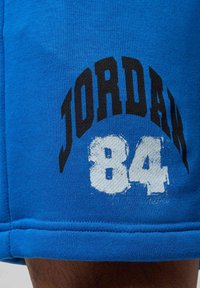 Niebieska tkanina z czarnym napisem "JORDAN" wygiętym nad wypłowiałymi białymi cyframi "84" nadrukowanymi na ubraniu w pobliżu dołu.