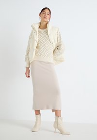 Pull en tricot beige avec un motif en losanges, gilet matelassé crème et jupe midi beige clair, associés à des bottines à talons avec fermeture éclair.