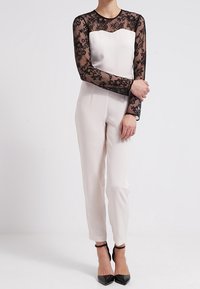 White jumpsuit with long black lace sleeves. Fitted bodice, tapered pants, features a heart-shaped neckline and a smooth texture. Black heels.