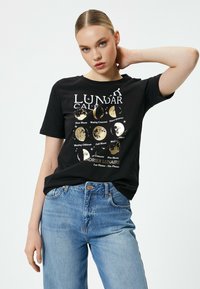 Sort bomulds T-shirt med grafik af månens faser i guld. Rund hals, korte ærmer, afslappet pasform, parret med højtaljede blå jeans.