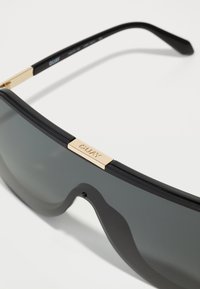 Zwarte zonnebril met een gebogen lens, gouden metalen accent bij de brug en slanke zwarte pootjes. Gemosse rand rond de lens.