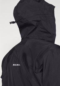 Czarna wodoodporna kurtka z kapturem, o gładkiej fakturze i minimalistycznym designie. Logo "MAMMUT" haftowane na boku w kolorze białym.