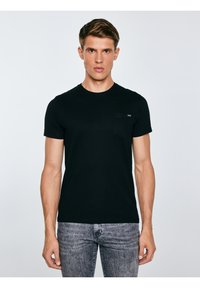 Noor mees lühikeste pruunide juustega, kes kannab musta lühikese varrukaga särk ja halli teksapükse, seisab tavalise heleda tausta ees.