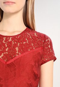 Haut en dentelle rouge avec un motif floral, à manches courtes, un col rond et une partie inférieure rouge unie, agrémentée d'une finition en satin.