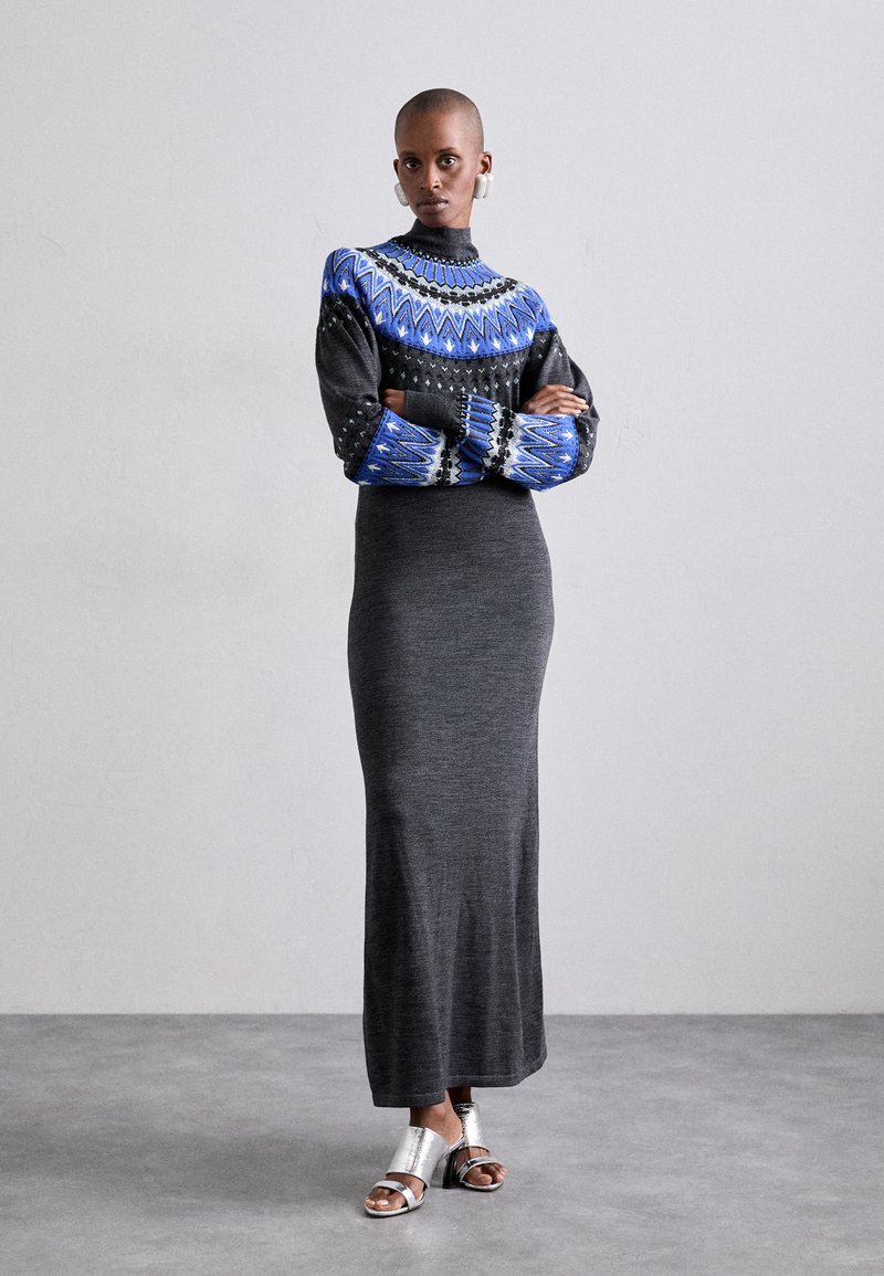 Rochie tricotată neagră cu mâneci lungi, top colorat cu model și guler înalt. Modelul poartă sandale argintii cu un finisaj metalizat.