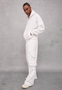 Sudadera blanca con cremallera frontal, bolsillos laterales y puños acanalados, combinada con pantalones cargo de corte relajado a juego. Textura del tejido suave en todo el conjunto.