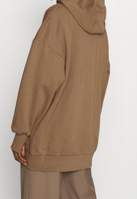 Felpa con cappuccio oversize di un colore marrone tenue, realizzata in tessuto morbido. Presenta spalle scese e polsini a coste, con una vestibilità comoda.