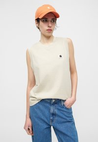 Γυναίκα που φοράει μπεζ αμάνικη μπλούζα με λογότυπο, μπλε τζιν και πορτοκαλί καπέλο, στέκεται με το ένα χέρι στην τσέπη, μπροστά σε λευκό φόντο.