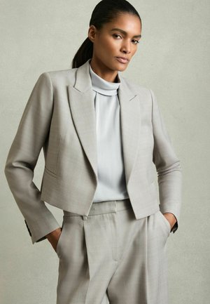 Světle šedý zkrácený blazer s jemným vzorem herringbone, v kombinaci s hladkým svetrem se stojáčkem ve světle šedé barvě. Přiléhavý střih se strukturovanými rameny.