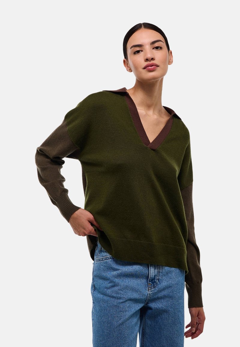 Pull-over en V vert et marron avec des manches longues, présentant un col contrasté et un tissu texturé. Associé à un jean en denim bleu.