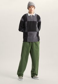 Ung person som bär grå mössa, svart och grå lapptäcks-långärmad skjorta, lösa gröna byxor och svartvita rutmönstrade slip-ons.