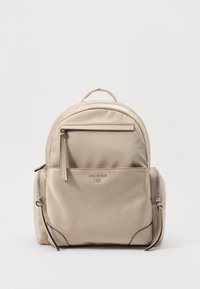 Zaino in nylon beige con forma arrotondata. Presenta una tasca frontale con zip, scomparti laterali con zip e una maniglia superiore. Dettaglio del logo metallico.