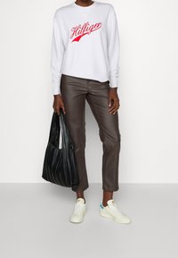 Sudadera blanca con texto rojo "Hilfiger", pantalones de cuero marrón, bolsa negra con pliegues y zapatillas blancas con acento turquesa.