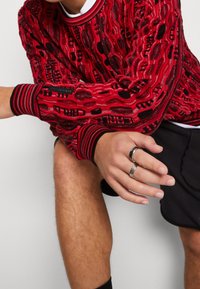 Rode textuurtrui met zwarte accenten, voorzien van een patroonontwerp. Geribbelde manchetten en halslijn, gedragen met zwarte shorts en metalen ringen.
