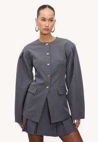 Blazer gris sur mesure avec fermetures à boutons argentés, jupe plissée et épaules structurées. Comprend deux poches avant et une texture lisse.