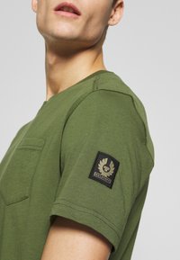 Man med olivgrön t-shirt med bröstficka och svart Belstaff-logotyp på vänster övre ärm mot en enhetlig bakgrund.